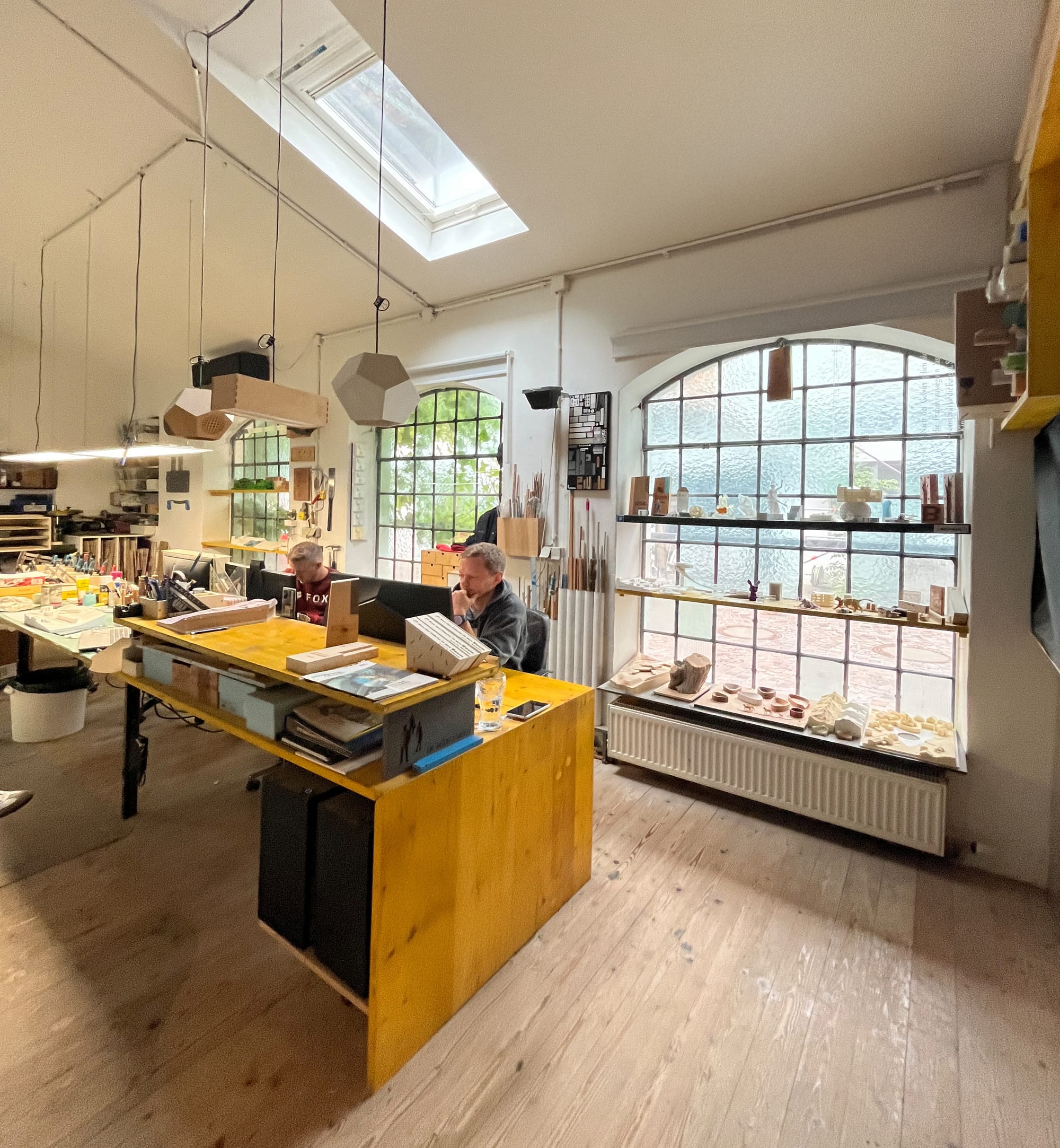 Die Atelierwerkstatt von innen: langer Holztisch, Rundbogenfenster und Werkzeug an den Wänden.