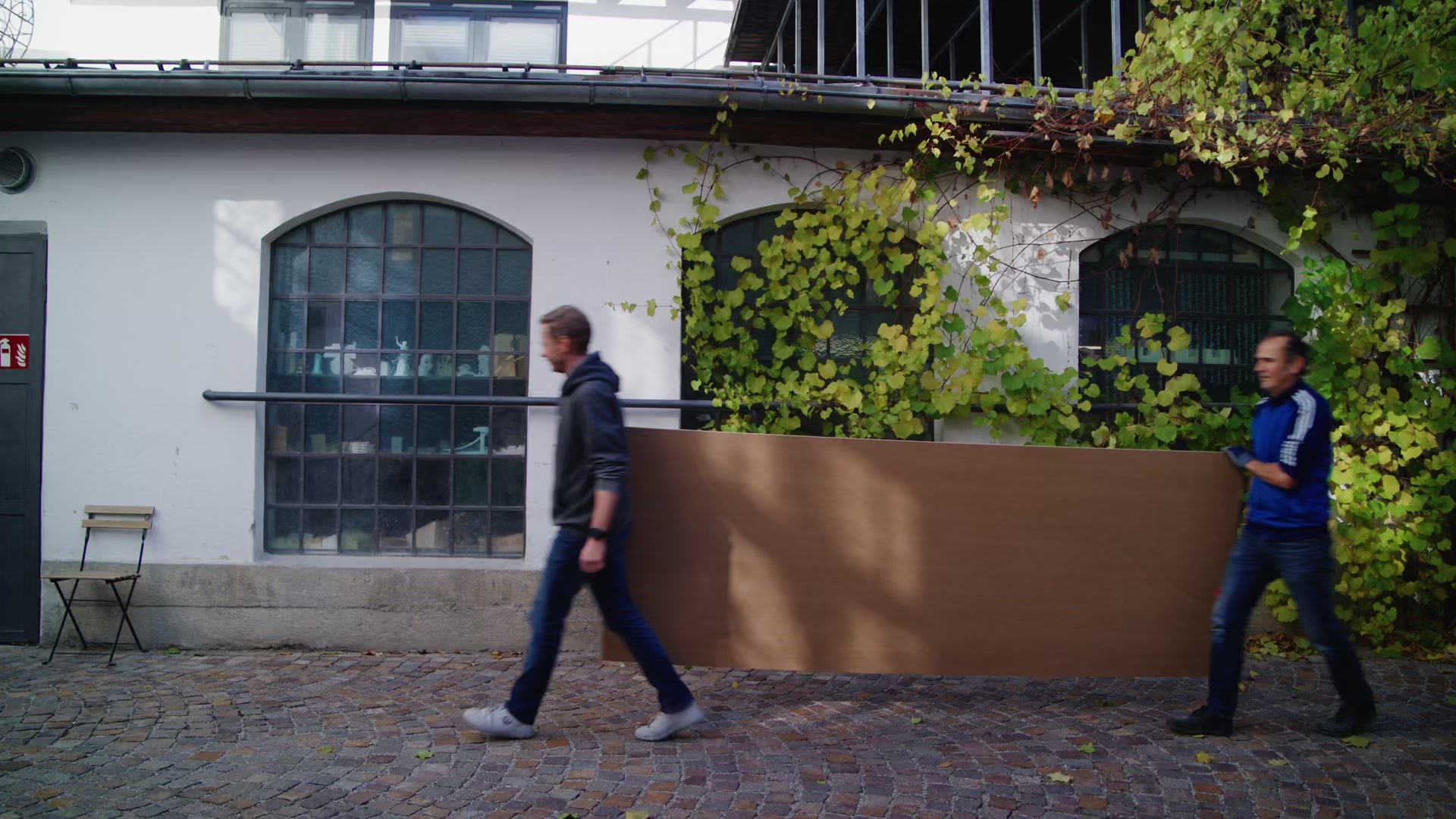 Zwei Modellbauer tragen eine große Holzplatte durch den begrünten Innenhof der Werkstatt.